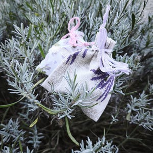 Könnte beinhalten: Zwei kleine Stoffsäckchen mit Lavendelzweig-Aufdruck. Das vordere Säckchen ist beige mit violetten Lavendelmustern und einem hellvioletten Band. Das hintere Säckchen ist weiß mit kleinen violetten Herzen und einem rosa Garnverschluss.