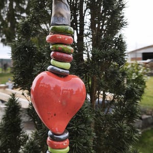 Könnte beinhalten: Dekorativer Gartenstecker mit rotem Herz, gestapelten Keramikringen in Grün, Rot und Grau sowie einer kleinen, abstrakten Figur an der Spitze. Der Stecker ist vor einem Hintergrund aus grünem Laub angebracht.