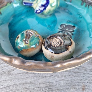 Könnte beinhalten: Zwei Keramikornamente in einer türkisfarbenen Schale. Ein Ornament ist grün und braun mit einem Wirbelmuster. Das andere Ornament ist schwarz und braun mit einem Blumenmuster.