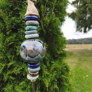 Könnte beinhalten: Ein Gartenstecker mit einer vertikalen Anordnung aus Keramikelementen, darunter ein beiges, spitz zulaufendes Oberteil, mehrere kleine blaue und graue Scheiben sowie eine große, zentrale, kugelförmige Perle mit glänzender, marmorierter Glasur.