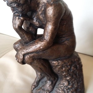 Op de afbeelding: Een bronzen sculptuur van een zittende figuur, met het hoofd op de hand. De figuur zit op een getextureerde basis. De sculptuur heeft een ruwe, verouderde uitstraling.