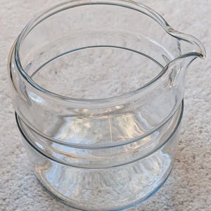 以下が含まれることがあります： 注ぎ口が付いた透明なガラスピッチャー。ピッチャーは、上部がフレアになった円筒形の本体と、ハンドルなしのデザインです。ガラスは透明で、内容物が見えます。ピッチャーはおそらく飲み物を提供するのに使用されます。