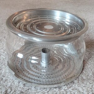 May include: A clear glass container with a cylindrical shape, featuring a removable metal lid and a metal base insert. Both metal components are circular with concentric rings of small perforations and a central vertical metal tube.