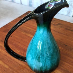 Peut inclure: Pichet en céramique avec une longue anse incurvée. Le haut et la poignée sont noirs, passant à un corps turquoise et vert. Le pichet a une finition brillante et repose sur une surface en bois.