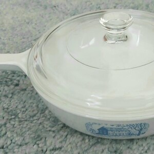 Puede incluir: Cazuela blanca con tapa de cristal transparente y asa blanca. El plato tiene una ilustración azul y blanca de una casa y árboles. El plato está sobre una superficie azul claro.