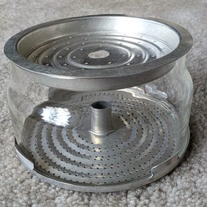 May include: A clear glass coffee pot with a metal filter and rim. The filter has concentric circles of small holes. The metal rim and filter are silver-coloured. The pot is designed for brewing coffee.