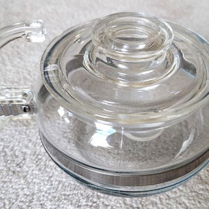 May include: A clear glass teapot with a matching lid and a curved handle. The teapot has a silver-coloured band around the middle and a spout for pouring. The teapot is designed for brewing tea.