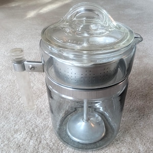 May include: A clear glass coffee percolator featuring a metal basket insert, a central metal stem with a base, and a glass lid with the text MADE IN U.S.A. embossed on the rim. The handle is made of clear plastic with a metal attachment bracket.