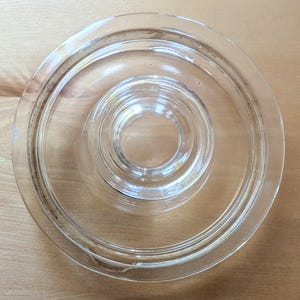 Puede incluir: Una tapa de vidrio transparente y circular con un diseño concéntrico escalonado y un asa central circular empotrada. La tapa tiene una superficie lisa y transparente con un borde exterior elevado, sirviendo como cubierta funcional.
