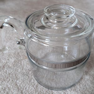 May include: A clear glass teapot with a matching glass lid and a side handle. The body of the teapot features a horizontal metallic band with a textured, ribbed pattern. The handle is attached to the main body with a silver-toned metal bracket.