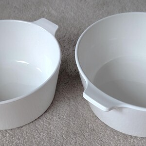Könnte beinhalten: Zwei weiße, runde Servierschalen mit Griffen. Die Schalen bestehen aus einem glatten, glänzenden Material und haben ein schlichtes, modernes Design. Die Schalen sind leer und scheinen sauber zu sein. Sie stehen auf einer hellgrauen Oberfläche.