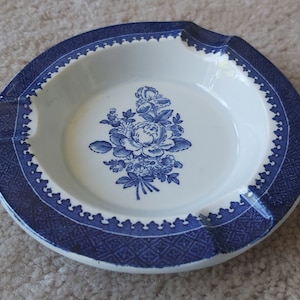May include: A circular, white ceramic ashtray with a blue floral pattern in the centre. The rim has a blue patterned border and a scalloped edge. It features three cigarette rests.