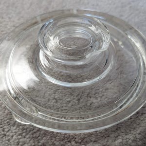 May include: A clear glass lid with a central, raised handle. The lid is circular with a slightly curved edge and a series of concentric rings forming the handle. The glass is transparent, allowing light to pass through.