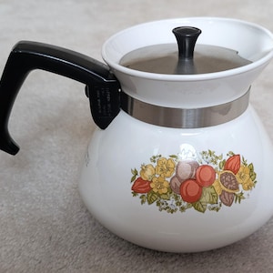 Könnte beinhalten: Weiße Emaille-Kaffeekanne mit schwarzem Griff und Deckel. Die Kanne hat ein dekoratives Blumen- und Fruchtdesign in Orange-, Gelb- und Brauntönen. Ein silbernes Band umschließt die Mitte der Kanne.