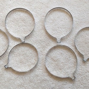 May include: Six stainless steel balloon-shaped moulds, each with a circular form and a small handle. The moulds are arranged on a light-coloured, textured surface. These are likely used for baking or cooking.