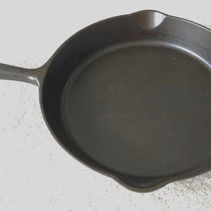 May include: A black cast iron frying pan with a long handle and a rounded cooking surface. The pan has a small spout on one side and a hole in the handle for hanging. The pan is designed for cooking.