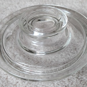 Puede incluir: Una tapa de vidrio transparente con diseño circular. La tapa presenta un centro elevado y redondeado, y un borde ancho y plano. El vidrio es transparente, permitiendo ver la superficie debajo. Diseño simple y funcional.