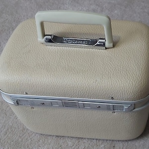 Puede incluir: Una maleta de viaje vintage de color crema con un exterior texturizado. Cuenta con un asa blanca y detalles metálicos plateados. La maleta tiene forma rectangular con esquinas redondeadas y un cierre seguro.