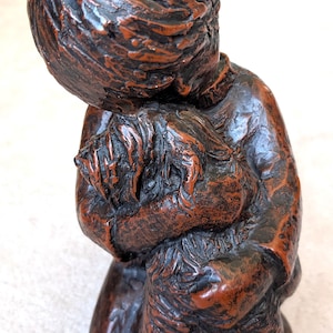 Op de afbeelding: Gedetailleerde sculptuur van een persoon die knielt en een andere figuur omarmt. Het beeld is afgewerkt in een donkerbruin en roodbruin kleurenschema, met gedetailleerde texturen. De figuren omarmen elkaar dicht.