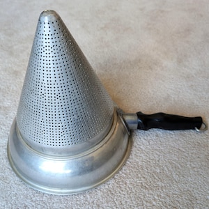 Puede incluir: Un pasapurés de metal vintage con un colador en forma de cono y un mango negro. El colador está perforado con numerosos agujeros pequeños. El pasapurés tiene una base ancha y poco profunda y un mango resistente. Herramienta de cocina para colar y hacer purés.