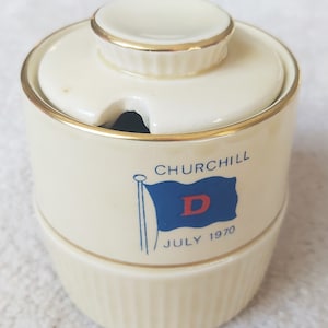 May include: Cream-coloured ceramic jar with a lid and gold trim. The jar features a blue flag with a red "D" and the text "CHURCHILL JULY 1970". The jar has a ribbed base and a small opening in the lid.