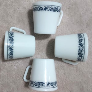 Puede incluir: Cuatro tazas de cerámica blanca con un diseño floral azul en el borde. Cada taza tiene un asa y una forma ligeramente acampanada. Las tazas están dispuestas en un patrón circular.