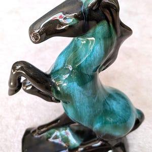May include: A ceramic horse figurine in a rearing pose. The horse has a glossy finish with a gradient of teal and black. The figurine is mounted on a rectangular base, also black.