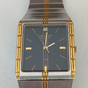 May include: A silver and gold two-tone watch with a square face and black dial. The watch has a gold band with a diamond at the 12 o'clock position. The watch face reads "Orient Quartz".