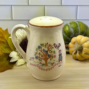 May include: Cream-colored ceramic salt shaker with a handle and a lid. The shaker is decorated with a tree, birds, a chicken, a girl, and a fence. The lid has three holes for dispensing salt. The shaker is a kitchenware item.