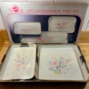 May include: A vintage 3-piece lacquerware tray set. The set includes three trays of varying sizes, each with a white background and a floral design in pink, blue, and yellow. The trays have gold-colored trim and black handles. The box reads "Pink Rose 3 PC. Lacquerware Tray Set."