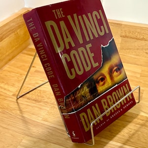 May include: A hardcover book titled "The Da Vinci Code" by Dan Brown, displayed in a silver metal book stand. The book has a burgundy cover with gold lettering and a partial image of a face. The book stand is on a wooden surface.