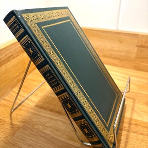 May include: A hardcover book titled "Captains Courageous" by Rudyard Kipling, displayed on a silver metal book stand. The book has a dark teal cover with gold decorative borders and lettering. The stand is made of thin, bent metal.