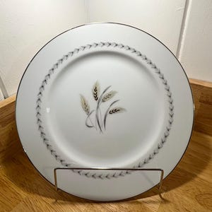 May include: White ceramic plate with a wheat stalk design. The plate has a silver rim and a decorative border of small, gray leaves. The wheat stalks are in shades of tan and gray. The plate is displayed on a metal stand.