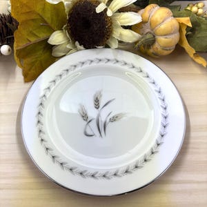May include: White oval plate with a silver rim and a wheat stalk design in the center. The plate is surrounded by a gray leaf pattern. The plate is on a light wood surface, with fall-themed decorations in the background.