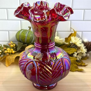 May include: A ruby red glass vase with a ruffled rim and iridescent finish. The vase features a bulbous body with embossed floral and geometric patterns. The vase is approximately 8 inches tall.