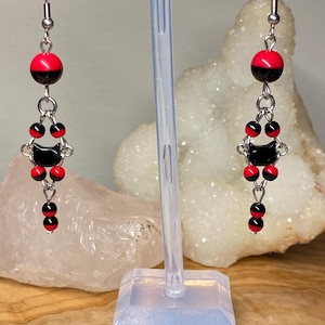 May include: A pair of dangle earrings featuring a black cat design. The earrings have red and black beads, with a silver-tone metal hook. The earrings are displayed on a clear stand against a blurred background.