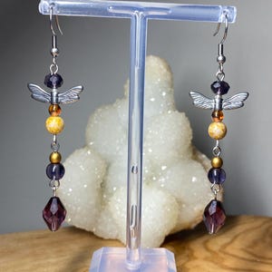 May include: A pair of dangle earrings with a silver-tone angel wing charm, purple and yellow beads, and a faceted purple teardrop. The earrings are displayed on a clear stand against a neutral background.