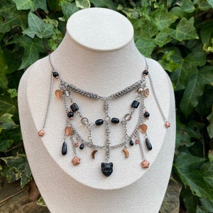 May include: A silver chain necklace with black and clear beads, copper stars, and crescent moons. The necklace has a black cat charm in the centre.