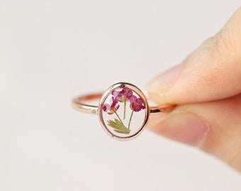 May include: A rose gold ring featuring an oval-shaped bezel setting that contains clear resin embedded with small, delicate pink flowers and green stems. The metal band is thin and polished, showcasing a botanical design within the center piece.