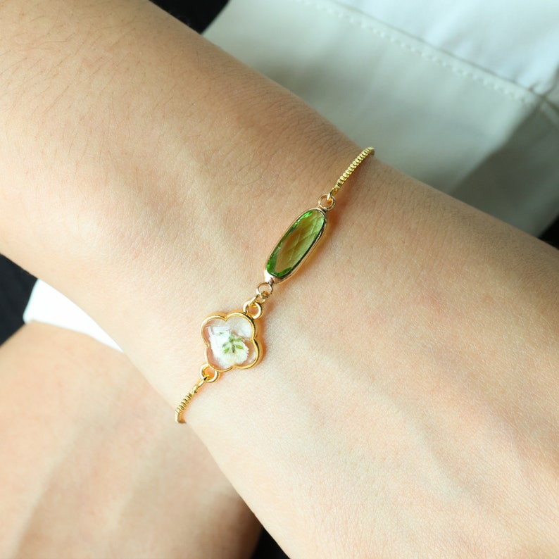 May include: A gold chain bracelet with a green oval-shaped gemstone and a clover-shaped charm with a white flower pressed inside.