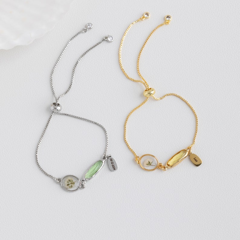 Puede incluir: Dos pulseras ajustables con cadenas delicadas y peque&ntilde;os colgantes ovalados. Una pulsera es plateada con un colgante de flor prensada verde y la otra es dorada con un colgante de flor prensada blanca. Ambos colgantes tienen una peque&ntilde;a etiqueta con texto.