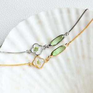 May include: Two delicate bracelets with a clover-shaped charm and a green oval-shaped stone. One bracelet has a silver chain and the other has a gold chain.