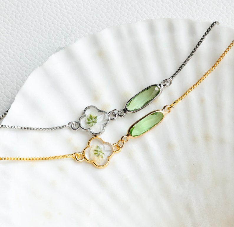 May include: Two delicate bracelets with a clover-shaped charm and a green oval-shaped stone. One bracelet has a silver chain and the other has a gold chain.