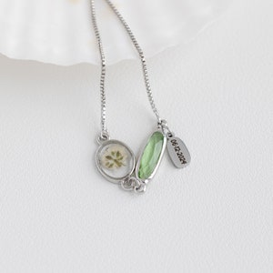 Puede incluir: Un collar de cadena de plata con un colgante ovalado verde y un colgante ovalado m&aacute;s peque&ntilde;o que contiene un tr&eacute;bol de cuatro hojas prensado. El colgante tiene una etiqueta de plata grabada con la fecha "06-12-2024".