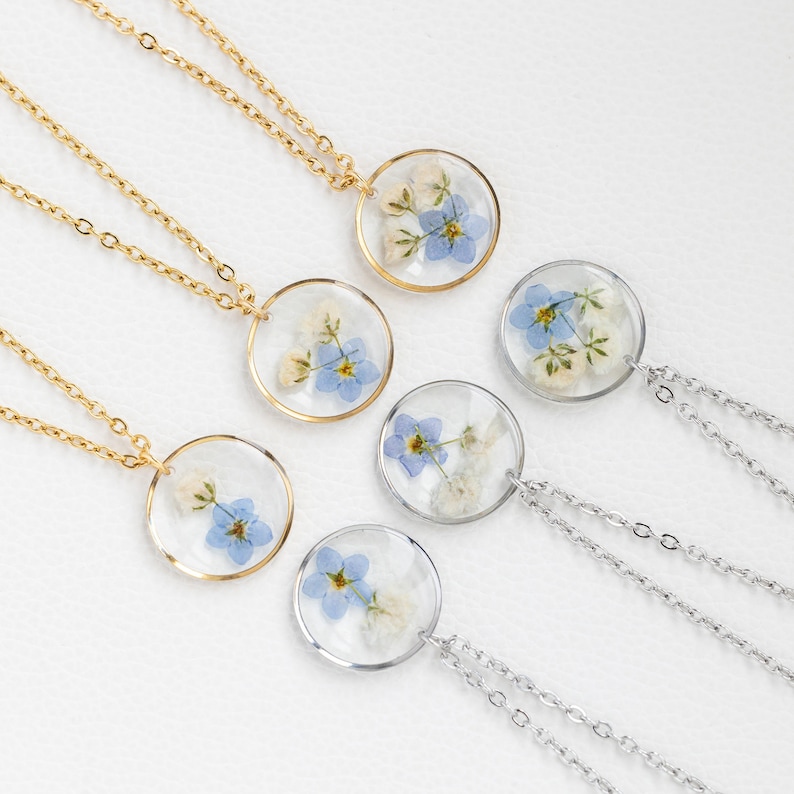 May include: Four necklaces with delicate silver and gold chains and round pendants featuring pressed forget-me-not flowers in clear resin.