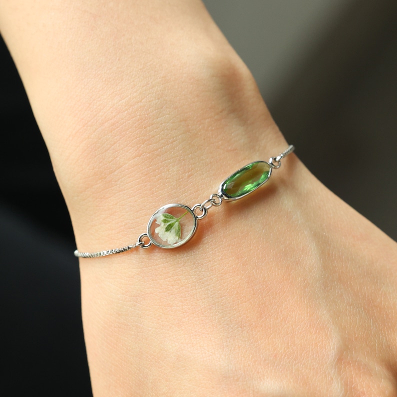 May include: A silver chain bracelet with two oval charms. One charm features a pressed green leaf, and the other charm features a pressed white flower.