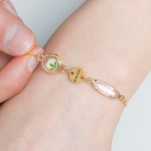 May include: Gold bracelet with a clear oval charm containing a pressed white flower, a round charm with the date "01.06.2023" engraved on it, and a pink oval charm.