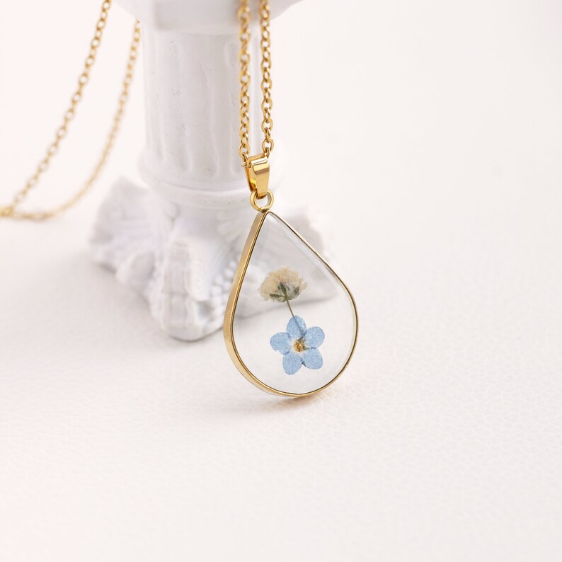 K&ouml;nnte beinhalten: Eine goldene Kettenkette mit einem tropfenf&ouml;rmigen Anh&auml;nger. Der Anh&auml;nger ist mit klarem Harz gef&uuml;llt und enth&auml;lt eine gepresste blaue Blume und eine kleine wei&szlig;e Blume.