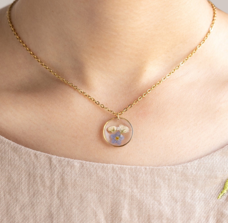 May include: A gold chain necklace with a round pendant featuring pressed blue and white flowers encased in clear resin.
