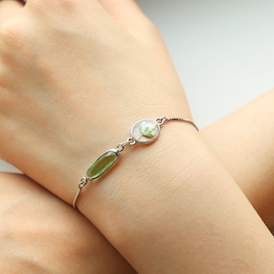May include: A silver bracelet with a green oval-shaped gemstone and a round clear charm with a pressed flower inside.